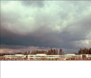 A dramatic cloudy sky looms over a cluster of white office buildings nestled between trees with autumn leaves. 